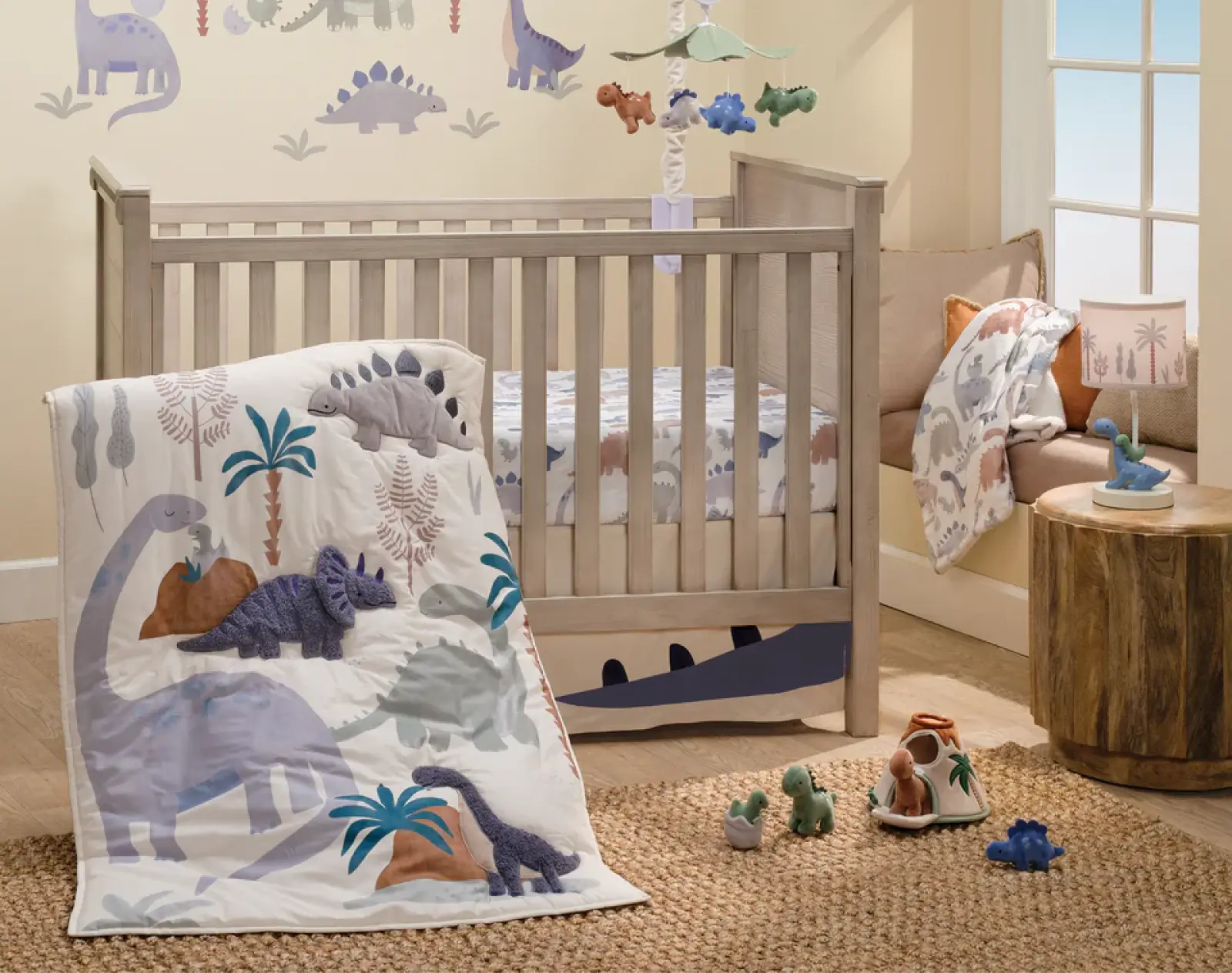 Neutral dinosaur crib bedding set with tropical palm tree and triceratops quilt, plush dinosaur toys, and wall decals in a beige nursery.